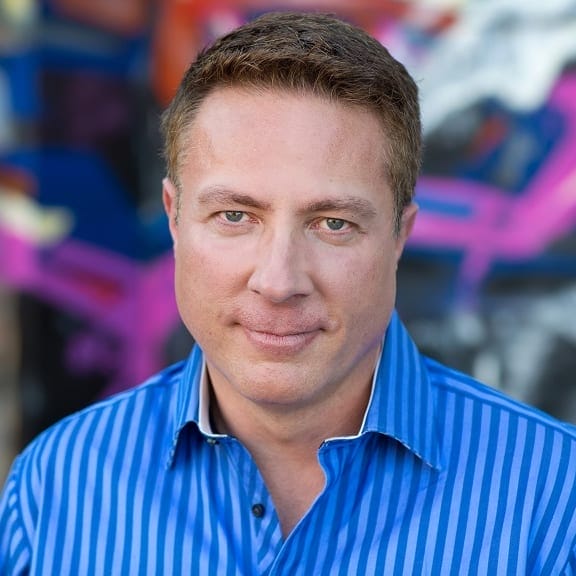 A man with short light brown hair wearing a blue striped shirt looks at the camera, standing in front of a colorful blurred background—perfect for Boston Band or wedding music promotional photos.