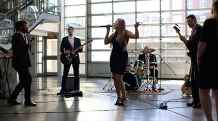 A Boston Band performs indoors with a female lead singer, guitarist, bassist, keyboardist, and drummer in a modern, well-lit space with large windows—perfect for unforgettable wedding music.