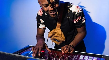 A man wearing a floral shirt and tan crossbody bag operates a DJ controller, smiling while mixing wedding music against a blue background.