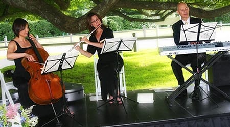 Three musicians from a Boston band perform wedding music outdoors with a cello, flute, and keyboard, each reading sheet music on stands under a tree.