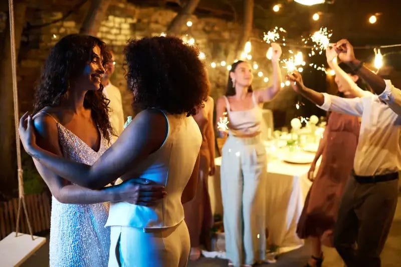 Two women embrace and dance at an outdoor party as friends celebrate with sparklers under string lights, enjoying lively wedding music performed by a Boston band.
