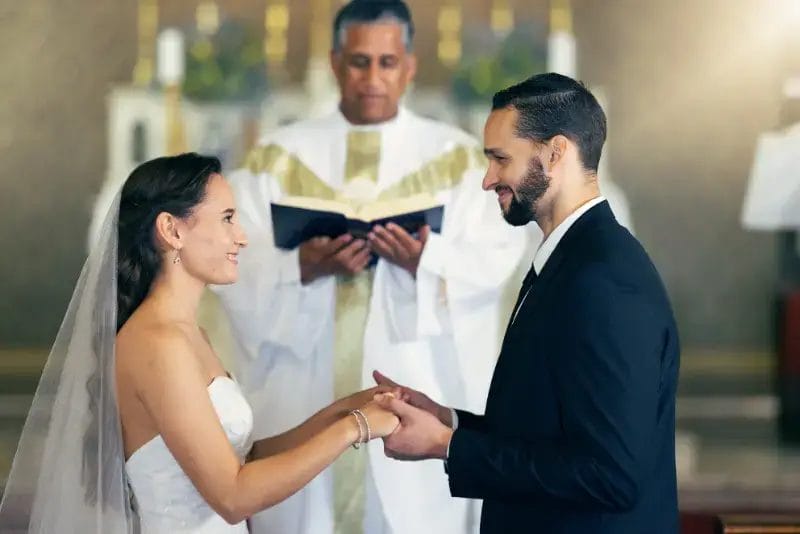 A bride and groom stand facing each other, holding hands during a wedding ceremony, while an officiant in robes reads from a book behind them as Boston Band plays romantic wedding music in the background.