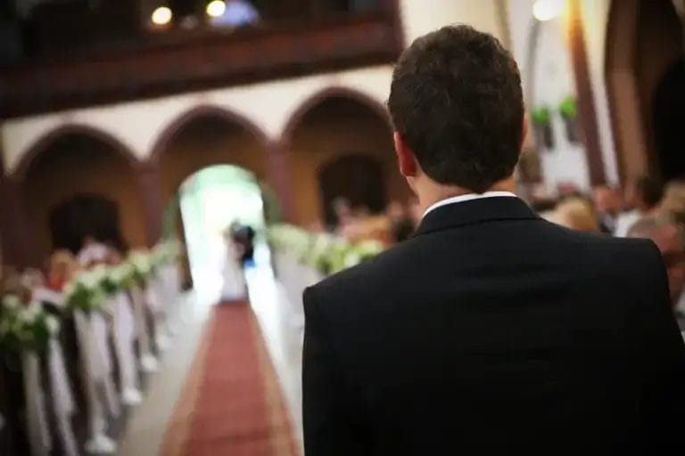 A man in a suit stands at the front of an aisle in a church, as a Boston Band softly plays wedding music while an approaching couple appears in the distance.