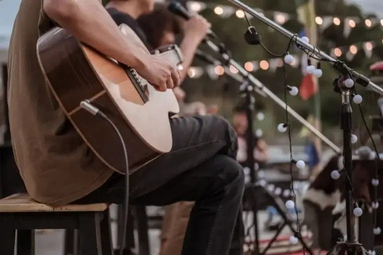 A person from a Boston Band plays acoustic guitar and sings into a microphone on an outdoor stage, string lights twinkling in the background—perfect for wedding music.