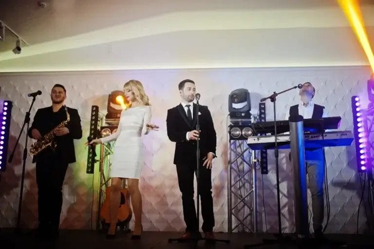 Four musicians from a Boston band perform on stage; one plays saxophone, one sings, one stands at a microphone, and one plays keyboards. Stage lights and instruments in the background create an energetic wedding music vibe.